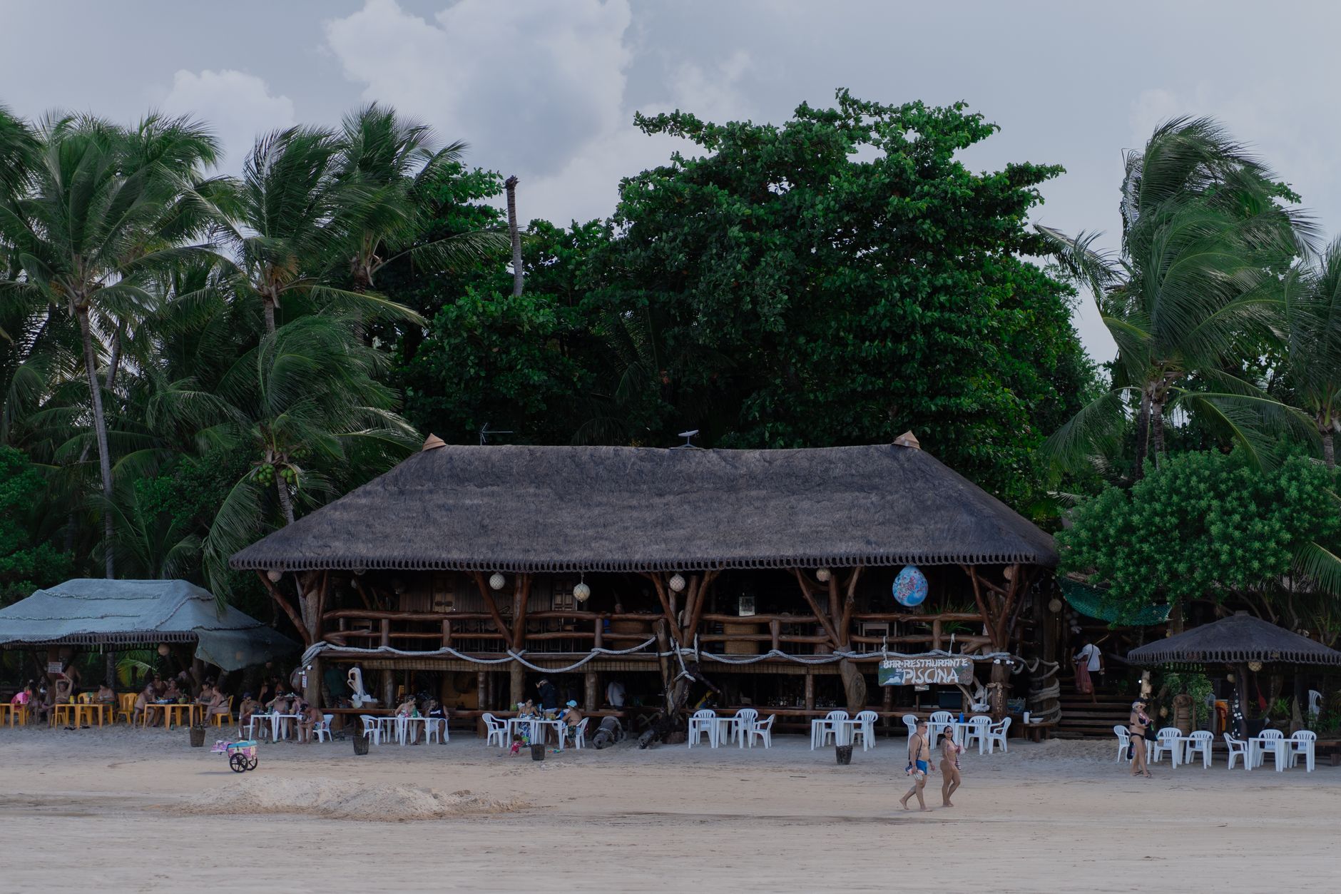 Rustic beachside restaurant with palm trees and people enjoying a summer day